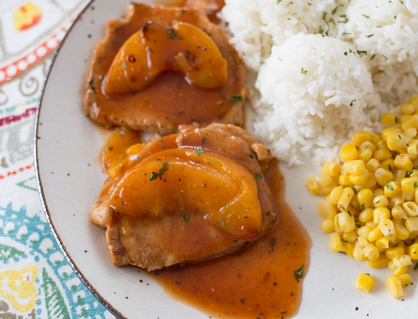close up photo of cooked Crock Pot Spicy Peach Pork Chops with a peach slice on each chop served on a glass dinner plate with cooked white rice and corn for the perfect side dish.
