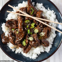 a dark blue bowl filled with tender Crock Pot Mongolian Beef served over a bed of cooked white rice with sliced green onions as the garnish.